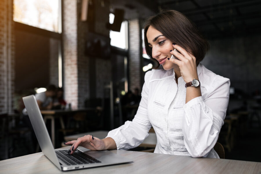 Mulher jovem em ambiente de escritório moderno, fazendo uma chamada telefônica enquanto trabalha em um laptop. Ideal para aprender como chamar paciente para retorno.