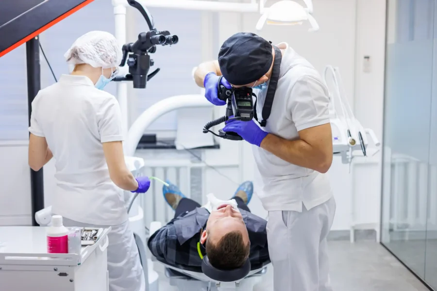 Fotografia odontológica em consultório, mostrando dentista registrando procedimento com câmera, auxiliado por um profissional, em ambiente clínico.